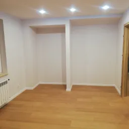 Pusty pokój po remoncie z drewnianą podłogą, białymi ścianami i wnękami, oświetlony punktowymi lampami sufitowymi.