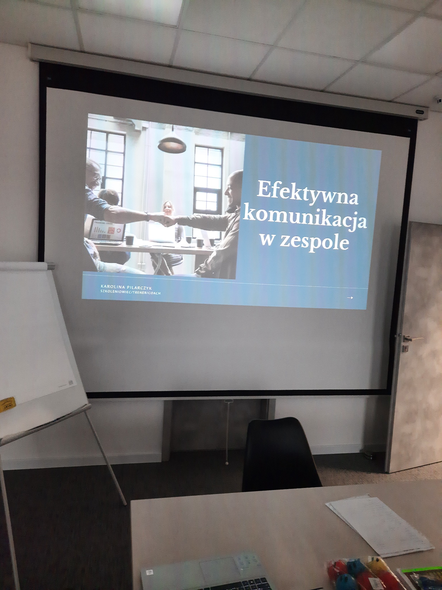 Prezentacja na ekranie projektora z napisem 'Efektywna komunikacja w zespole' i zdjęciem uścisku dłoni podczas spotkania biznesowego, widoczny flipchart i biurko z laptopem.