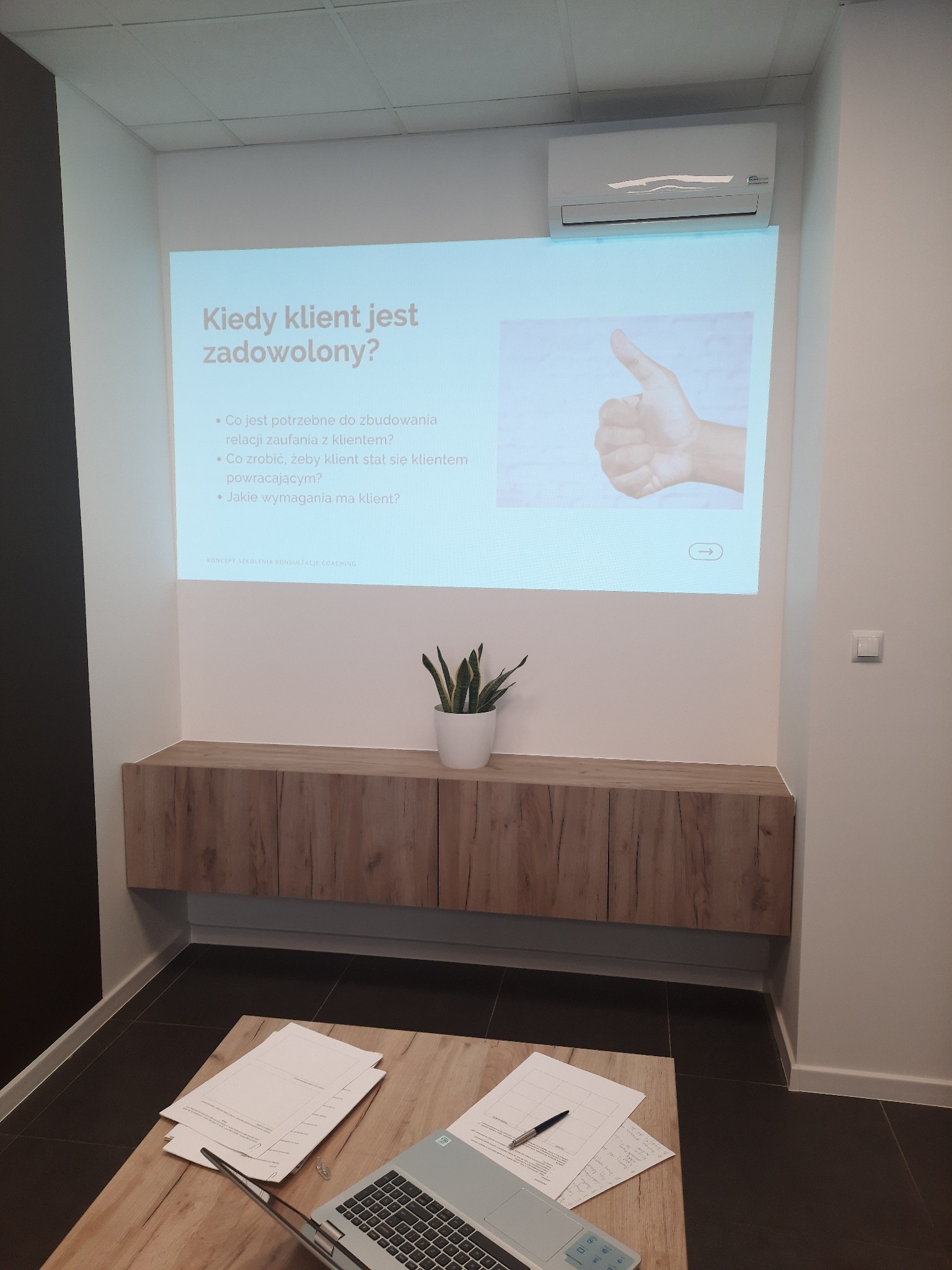Sala konferencyjna z projektorem wyświetlającym slajd prezentacji o budowaniu relacji z klientem, laptop i notatki na stole, szafka z rośliną, klimatyzator na ścianie.