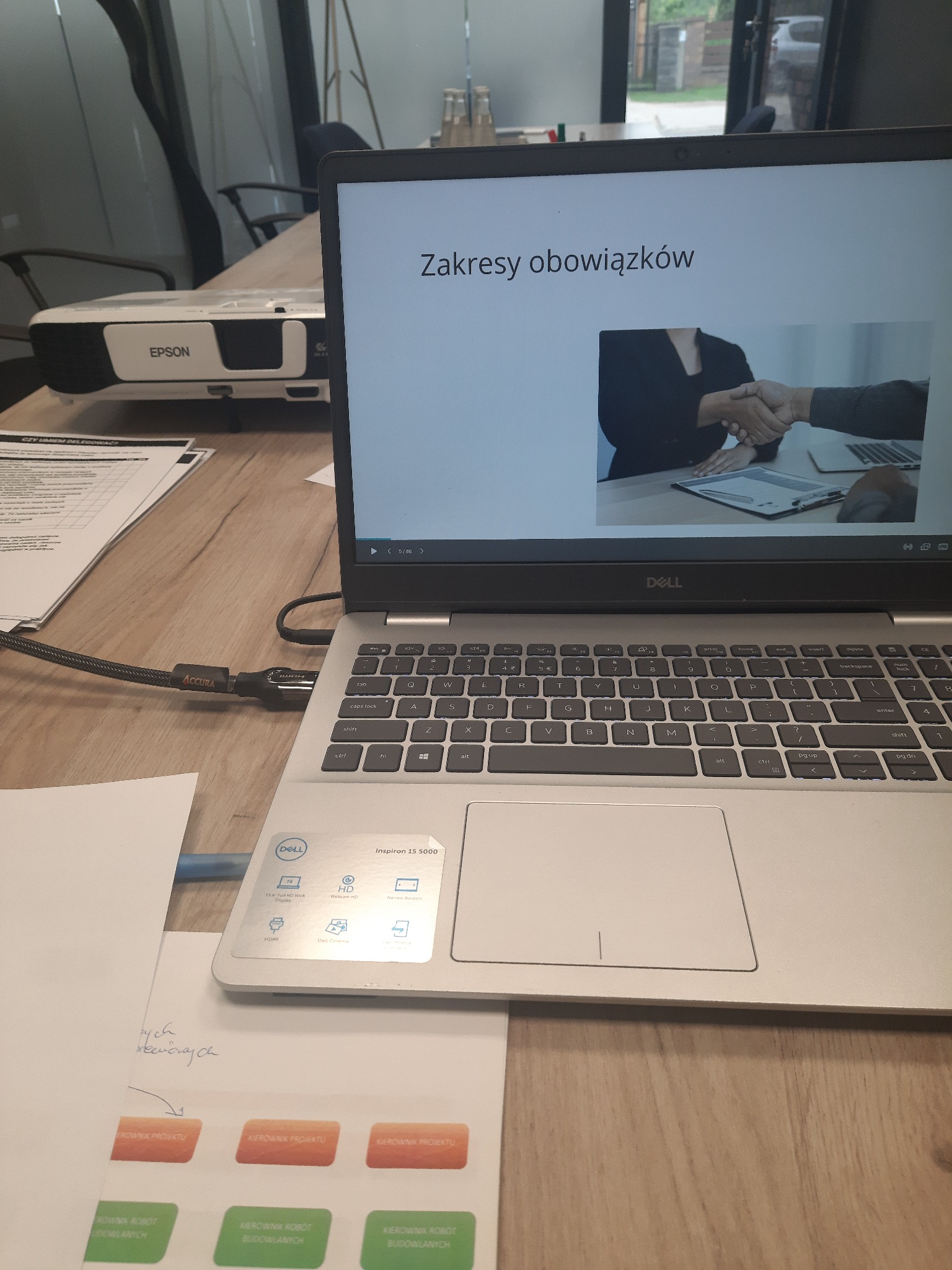Laptop Dell wyświetlający prezentację z nagłówkiem 'Zakresy obowiązków' i grafiką uścisku dłoni, obok leży projektor Epson, dokumenty z listą kontrolną i naklejki z napisem 'Kierownik Projektu'...