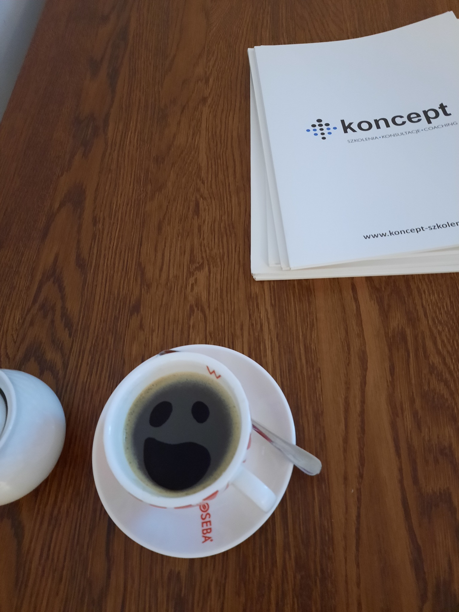 Filiżanka czarnej kawy z uśmiechniętą twarzą na powierzchni, obok cukiernica i stos białych kartek z logo 'koncept szkolenia konsultacje coaching' na drewnianym stole.