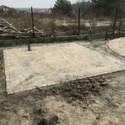 Przygotowany teren pod kostkę brukową z obrzeżami, widoczna rura kanalizacyjna i łopata oparta o ogrodzenie z siatki.