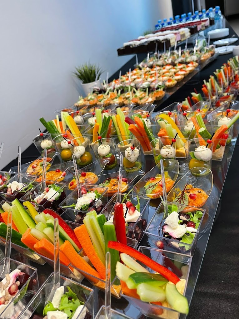 Długi stół cateringowy z przekąskami w małych, przezroczystych naczyniach: warzywa, sałatki i mini-kanapeczki. W tle woda i ciasta na paterach.