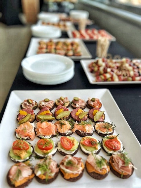 Elegancki catering: różnorodne, apetyczne kanapeczki na białych tacach, z widocznymi składnikami jak łosoś, pomidory i ogórki, ustawione na stole podczas eventu.