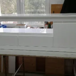 Biała, lakierowana szafka RTV z szufladami i otwartą półką, ustawiona na roboczym stole w warsztacie, widok z przodu na tle okna.