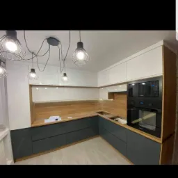 Nowoczesna kuchnia w kształcie litery L z szarymi szafkami dolnymi, białymi górnymi i drewnianym blatem, oświetlona wiszącymi lampami drucianymi.