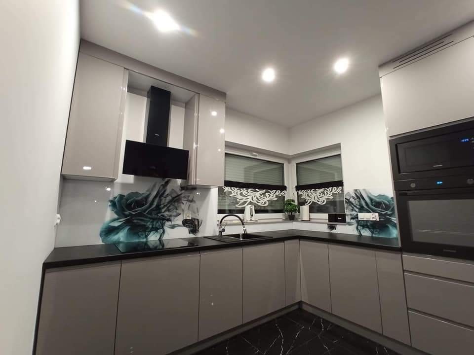 Nowoczesna kuchnia z szarymi szafkami, czarnym blatem i panelem szklanym z motywem kwiatowym, widok na okno z roletami.