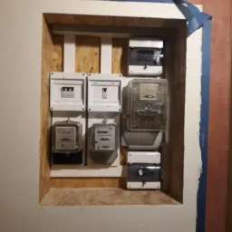 Szafka z licznikami i bezpiecznikami w ścianie, zabezpieczona niebieską taśmą malarską, widok z bliska.