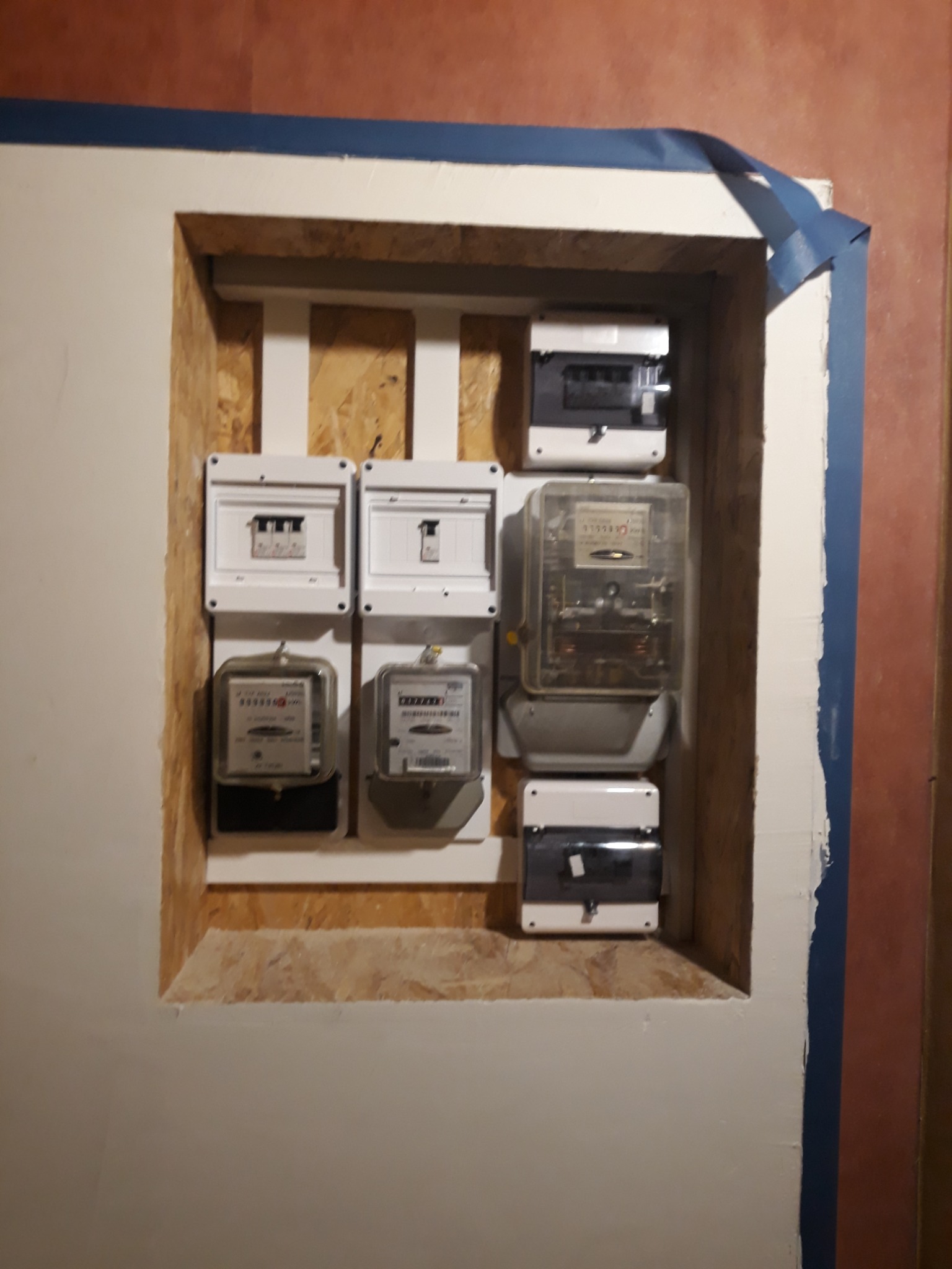 Szafka z licznikami i bezpiecznikami w ścianie, zabezpieczona niebieską taśmą malarską, widok z bliska.