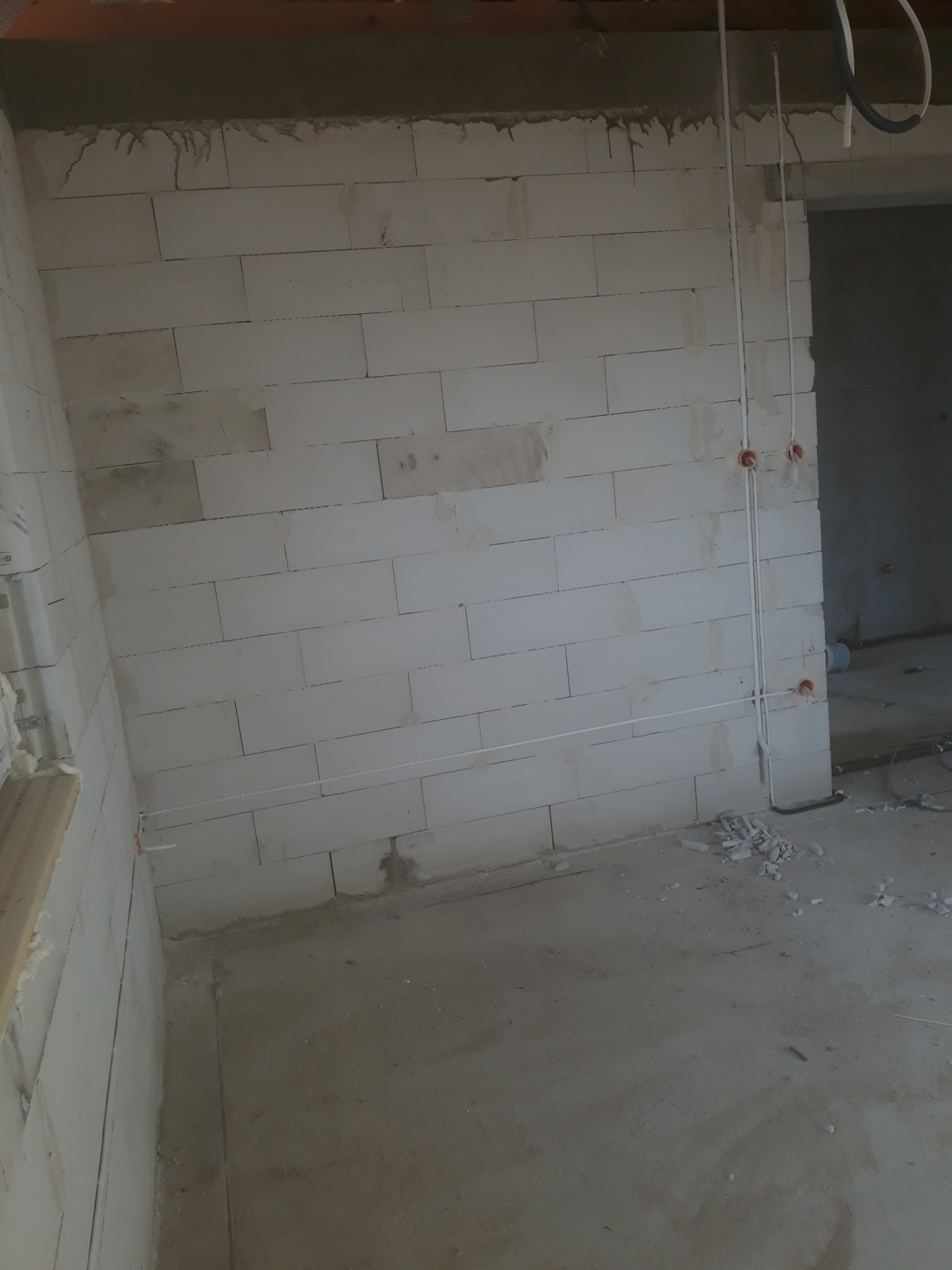 Wnętrze w trakcie prac wykończeniowych: ściany z białych bloczków, pionowe rury instalacji elektrycznej z widocznymi puszkami pod gniazdka, surowy beton na podłodze z resztkami gruzu.