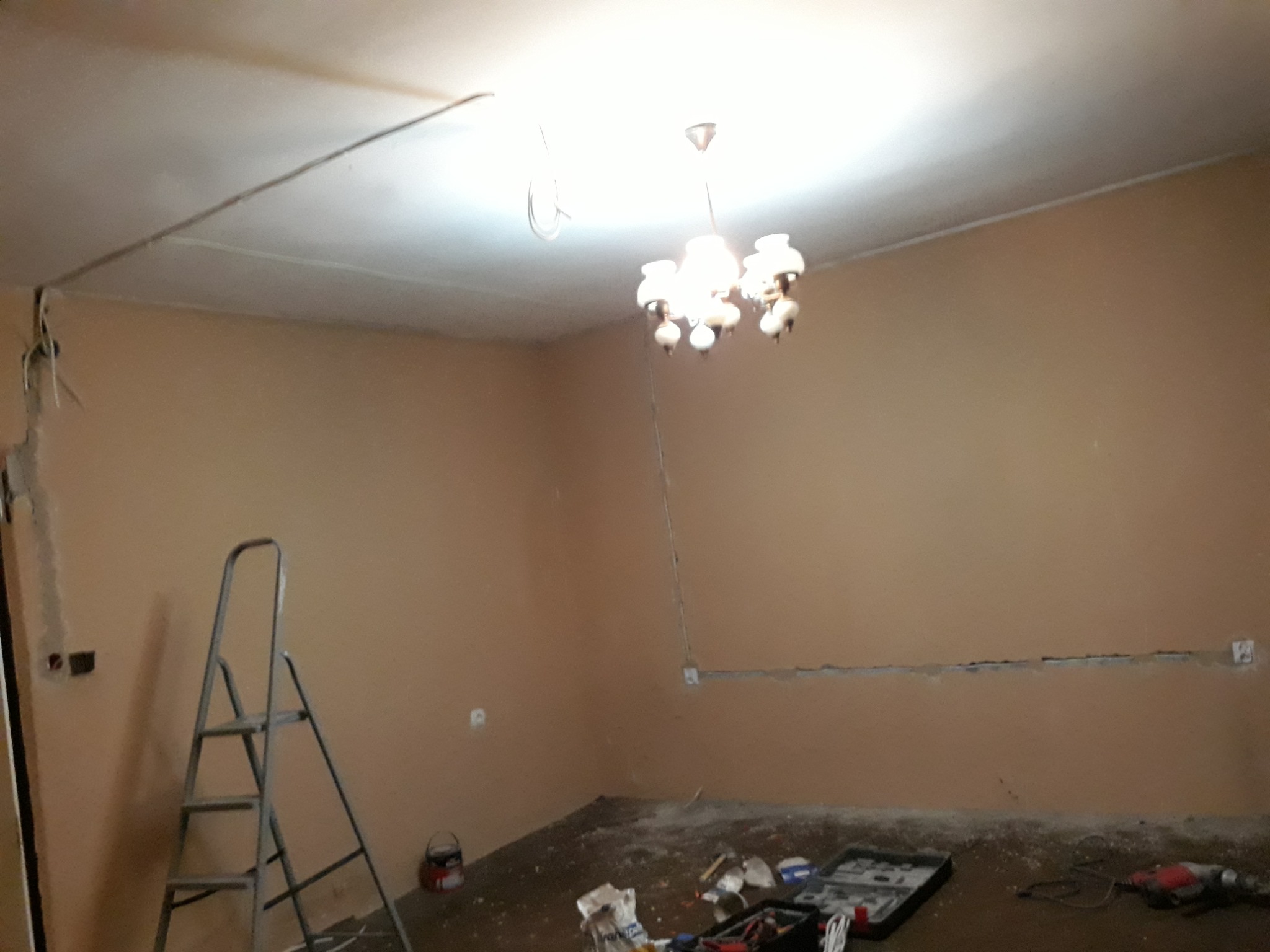 Pokój w trakcie remontu: widoczne ściany z bruzdami pod instalację elektryczną, drabina, narzędzia i wiszący żyrandol.