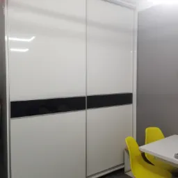 Biała szafa przesuwna z czarnym pasem dekoracyjnym, widok z boku, obok stoją dwa ż&oacute;łte krzesła i biały st&oacute;ł.