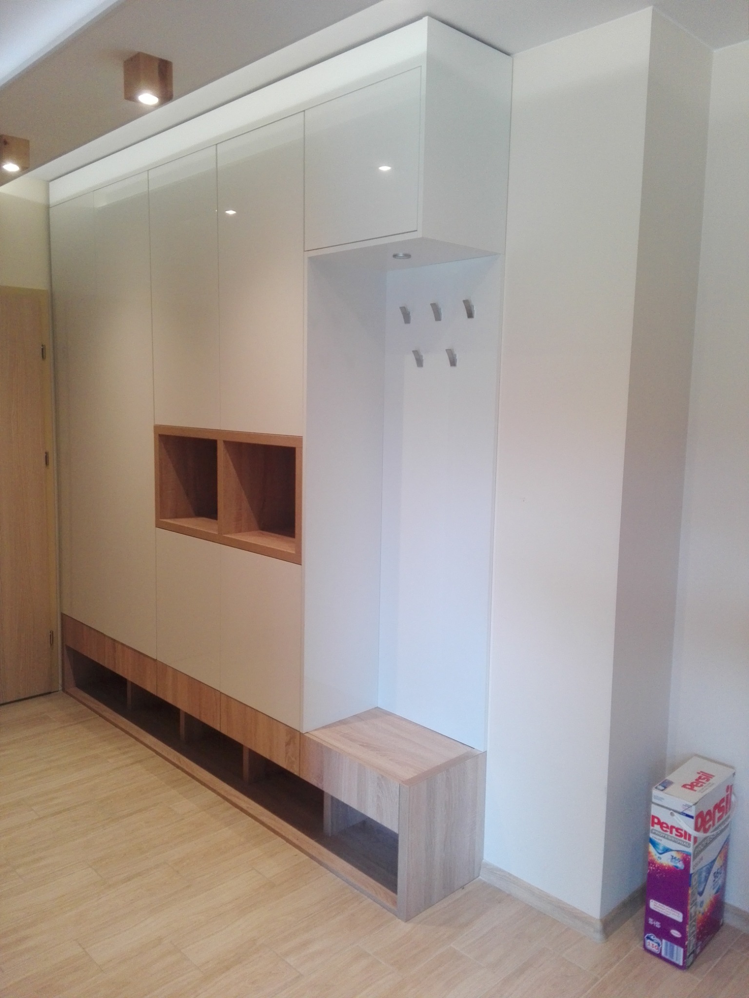 Jasna zabudowa przedpokoju z siedziskiem i wieszakami, uzupełniona o otwarte półki z drewnianym wykończeniem, widoczna pusta przestrzeń pod szafkami, na podłodze leży karton z proszkiem do prania.