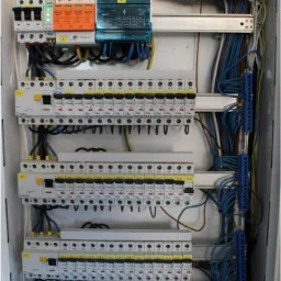 Otwarta skrzynka elektryczna z zainstalowanymi bezpiecznikami, ogranicznikami przepięć i licznymi przewodami. Widoczne listwy zaciskowe i oznaczenia na elementach. Na dole naklejka ostrzegawcza.