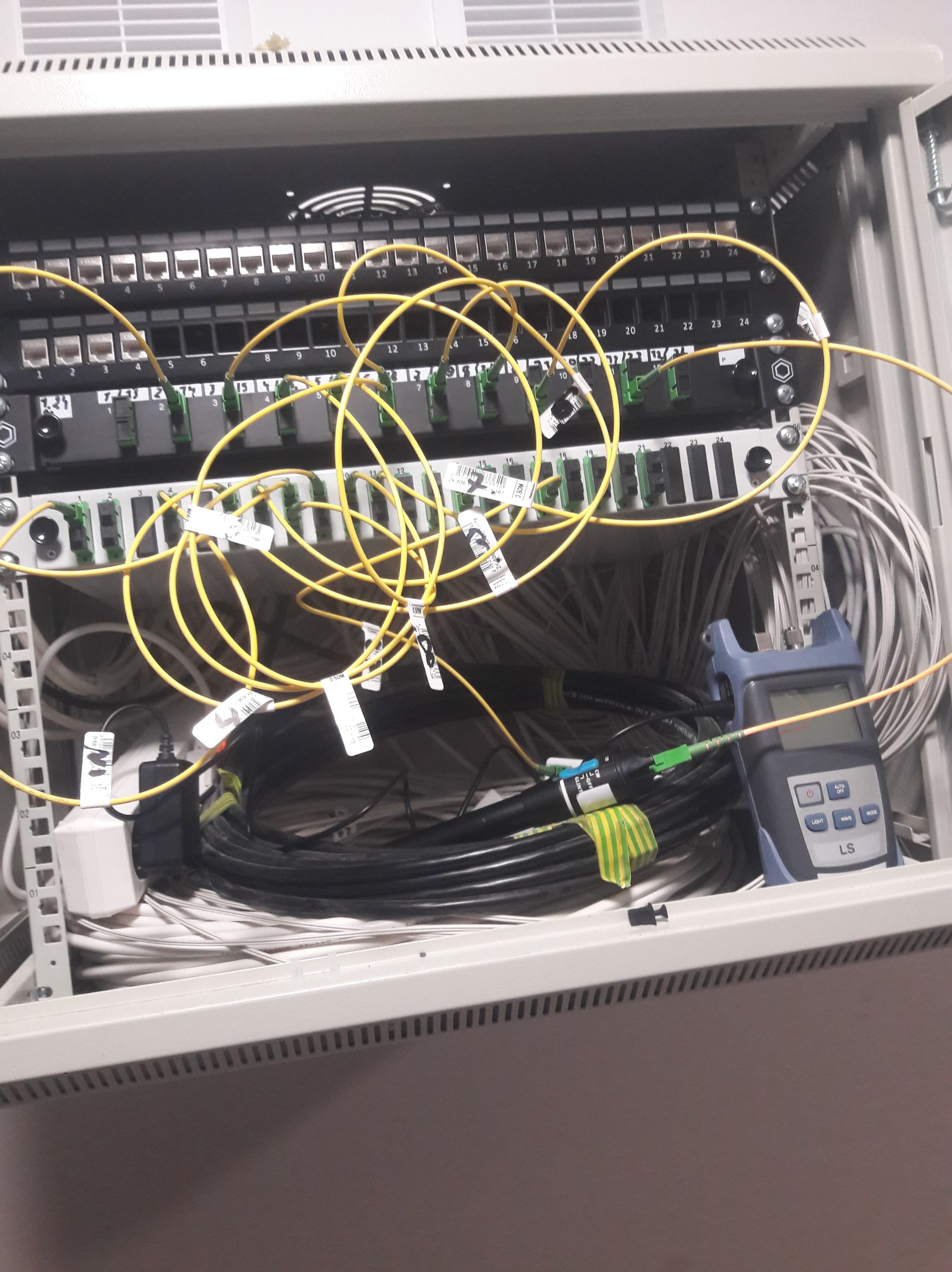 Panel krosowniczy światłowodowy w szafie rack z żółtymi patchcordami, miernikiem mocy optycznej LS i okablowaniem, widok z bliska.
