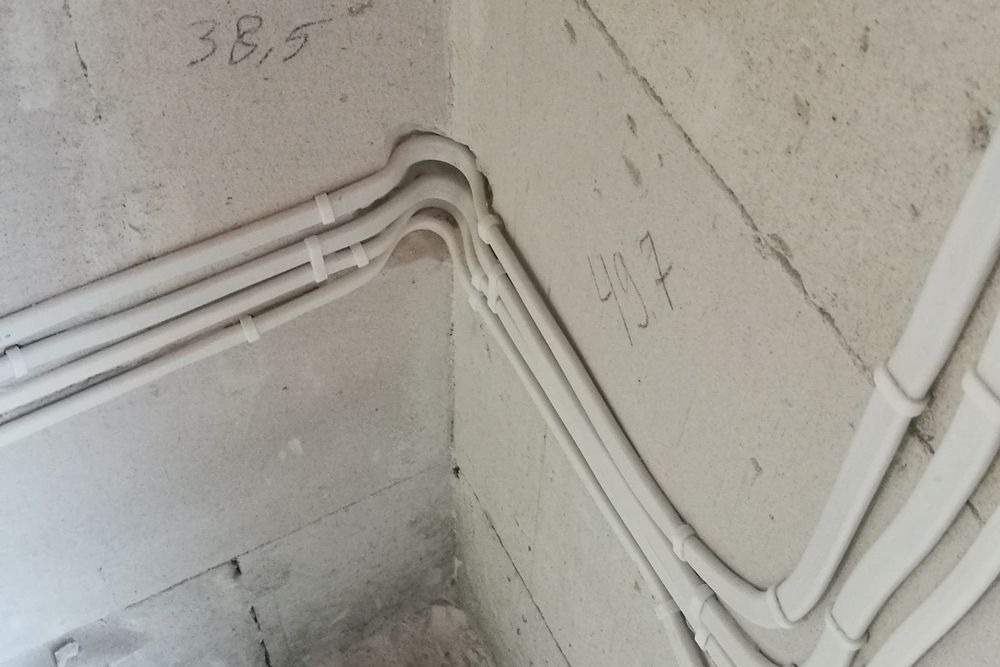 Białe kable elektryczne ułożone w narożniku surowej ściany z betonu komórkowego, przymocowane uchwytami, z odręcznymi notatkami '38,5' i '49.7' na ścianie.