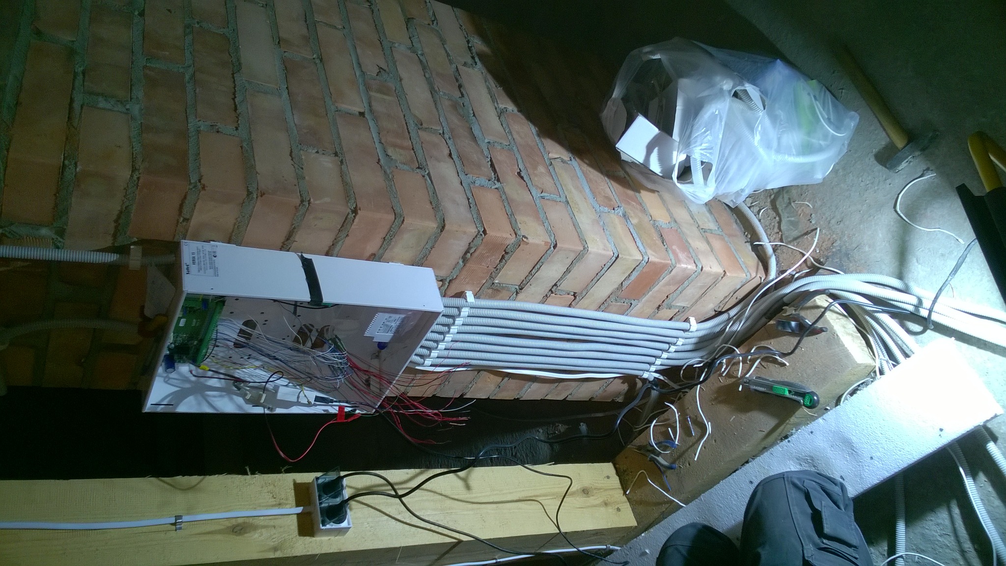 Rozdzielnica elektryczna z licznymi przewodami w peszlach zamontowana na ceglanej ścianie w piwnicy, widoczne narzędzia i elementy instalacji.