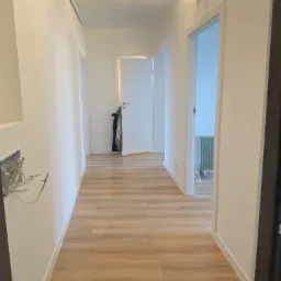 Korytarz po remoncie z panelami podłogowymi w odcieniach drewna, białe ściany i drzwi, widoczne niedokończone gniazdko elektryczne.