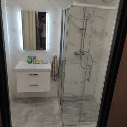 FIRMA REMONTOWO-BUDOWLANA PATBUD-REM PATRYK LECH - Nowoczesna łazienka z kabiną prysznicową typu walk-in, białą umywalką z szafką, lustrem z podświetleniem LED i marmurowymi płytkami na ścianach oraz szarymi płytkami podłogowymi; widoczne odbicie...