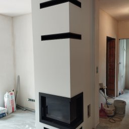 FIRMA REMONTOWO-BUDOWLANA PATBUD-REM PATRYK LECH - Biały narożny kominek z czarnymi akcentami w trakcie wykończenia, widoczne worki z zaprawą i narzędzia malarskie, surowy stan wykończenia pomieszczenia.