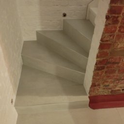 FIRMA REMONTOWO-BUDOWLANA PATBUD-REM PATRYK LECH - Betonowe schody w surowym wnętrzu z fragmentem ściany z czerwonej cegły, oświetlone z góry, widok z dołu.