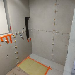 FIRMA REMONTOWO-BUDOWLANA PATBUD-REM PATRYK LECH - Ściany łazienki w trakcie układania płytek, widoczne listwy poziomujące i krzyżyki dystansowe, fragment podłogi zabezpieczony folią i taśmą malarską.