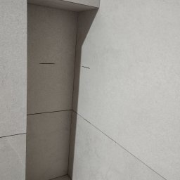 FIRMA REMONTOWO-BUDOWLANA PATBUD-REM PATRYK LECH - Minimalistyczne wykończenie narożnika łazienki z szarych, matowych płytek ceramicznych, z widocznymi fugami o delikatnej strukturze.