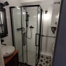 Nowoczesna kabina prysznicowa z czarnymi ramami i armaturą, białe płytki na ścianach i wzorzysta podłoga w łazience, umywalka na drewnianej szafce.