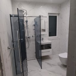 FIRMA REMONTOWO-BUDOWLANA PATBUD-REM PATRYK LECH - Nowoczesna łazienka z kabiną typu walk-in z przyciemnianego szkła, białą umywalką z szafką i toaletą. Marmurowe płytki na ścianach i podłodze. Minimalistyczny design.