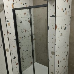 FIRMA REMONTOWO-BUDOWLANA PATBUD-REM PATRYK LECH - Kabina prysznicowa z czarnymi profilami na tle płytek lastryko w odcieniach bieli i beżu. Widoczny brodzik. Minimalistyczny design, nowoczesne wykończenie łazienki.