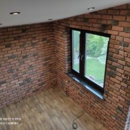 FIRMA REMONTOWO-BUDOWLANA PATBUD-REM PATRYK LECH - Pokój z ceglaną ścianą i drewnianą podłogą, widok z góry, czarna rama okienna z widokiem na zieleń, oświetlenie punktowe na suficie.