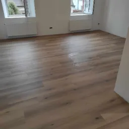 Ułożona podłoga z paneli drewnianych w jasnym odcieniu dębu w pustym pokoju z dwoma oknami i białymi ścianami, widoczne ślady po czyszczeniu na powierzchni.