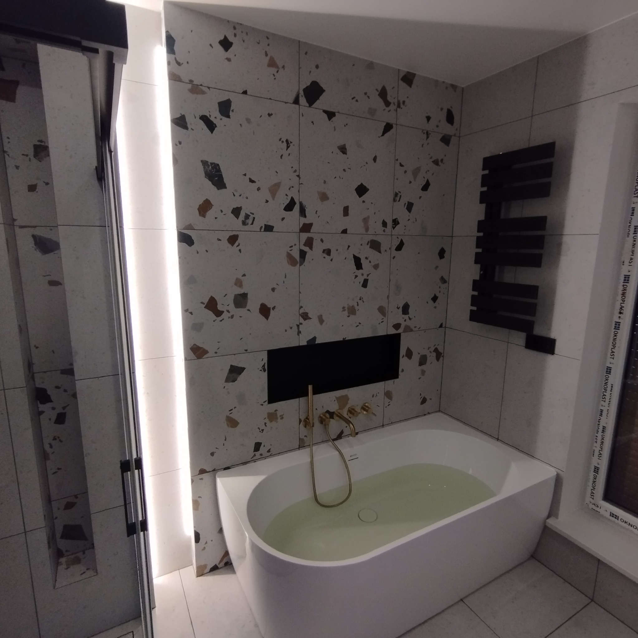 Biała wanna wypełniona wodą, złota bateria, ściany wyłożone płytkami w stylu lastryko, czarny grzejnik drabinkowy i wnęka w ścianie w wykończonej łazience.