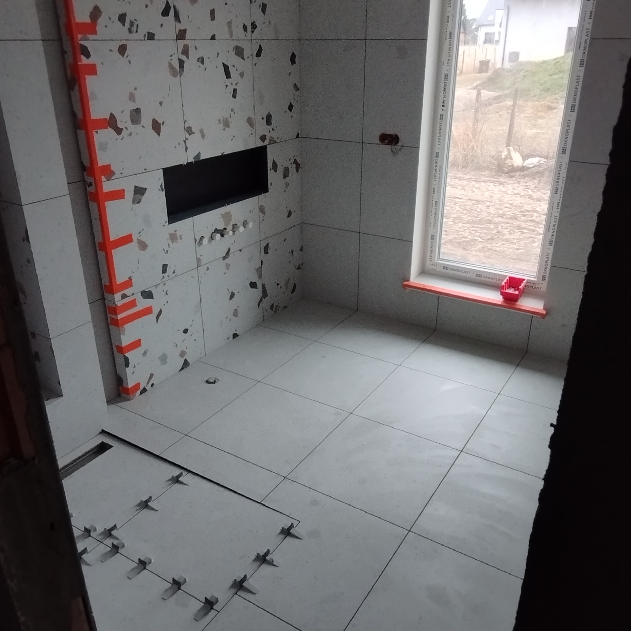 Łazienka w trakcie wykańczania, widoczne płytki imitujące lastryko na ścianach i podłodze, pomarańczowa taśma zabezpieczająca narożniki, poziomica przy ścianie i system poziomowania płytek...