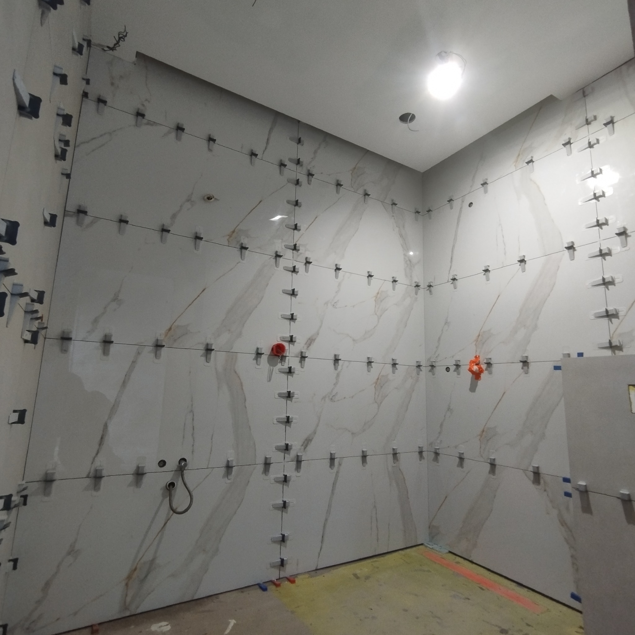 Ściany łazienki w trakcie układania płytek imitujących marmur, widoczne krzyżyki dystansowe i poziomowanie laserowe, instalacja wodna wystająca ze ściany.