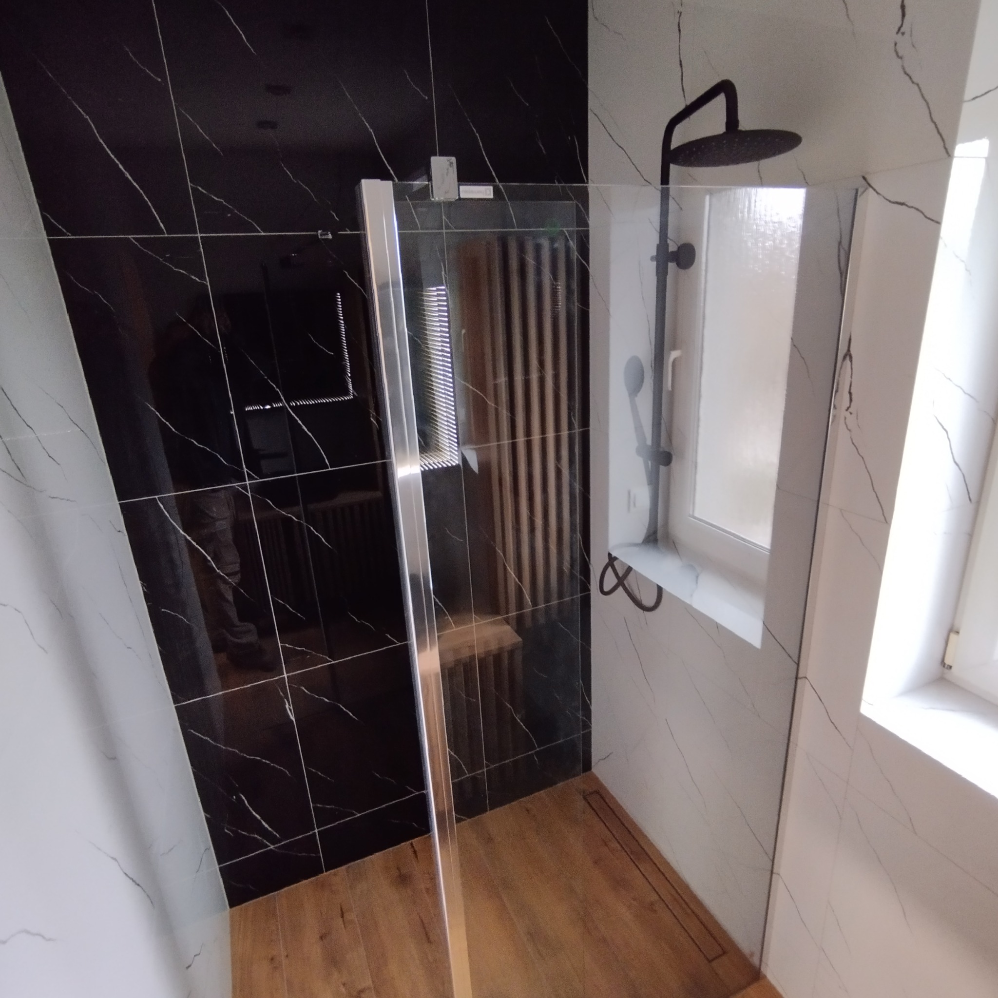 Nowoczesna kabina prysznicowa z czarnymi płytkami imitującymi marmur i drewnianą podłogą, widoczny odpływ liniowy i czarna deszczownica.
