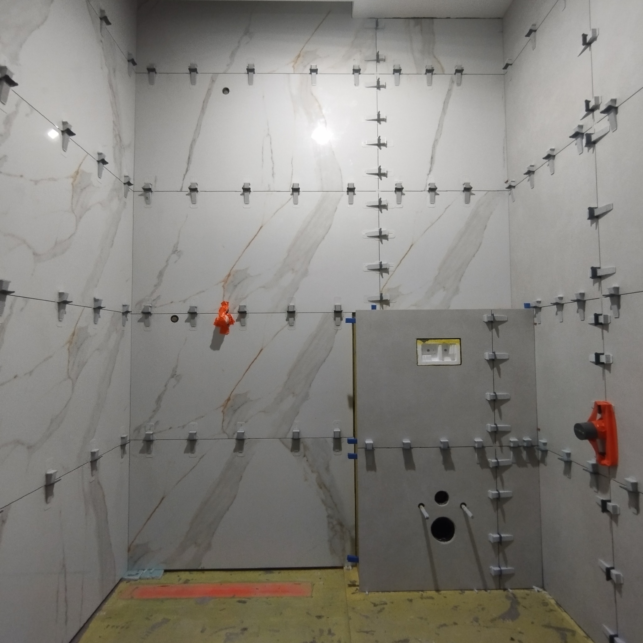 Ściany łazienki w trakcie układania płytek imitujących marmur, z zastosowaniem systemu poziomowania płytek; widoczny fragment podłogi z żółtą izolacją.