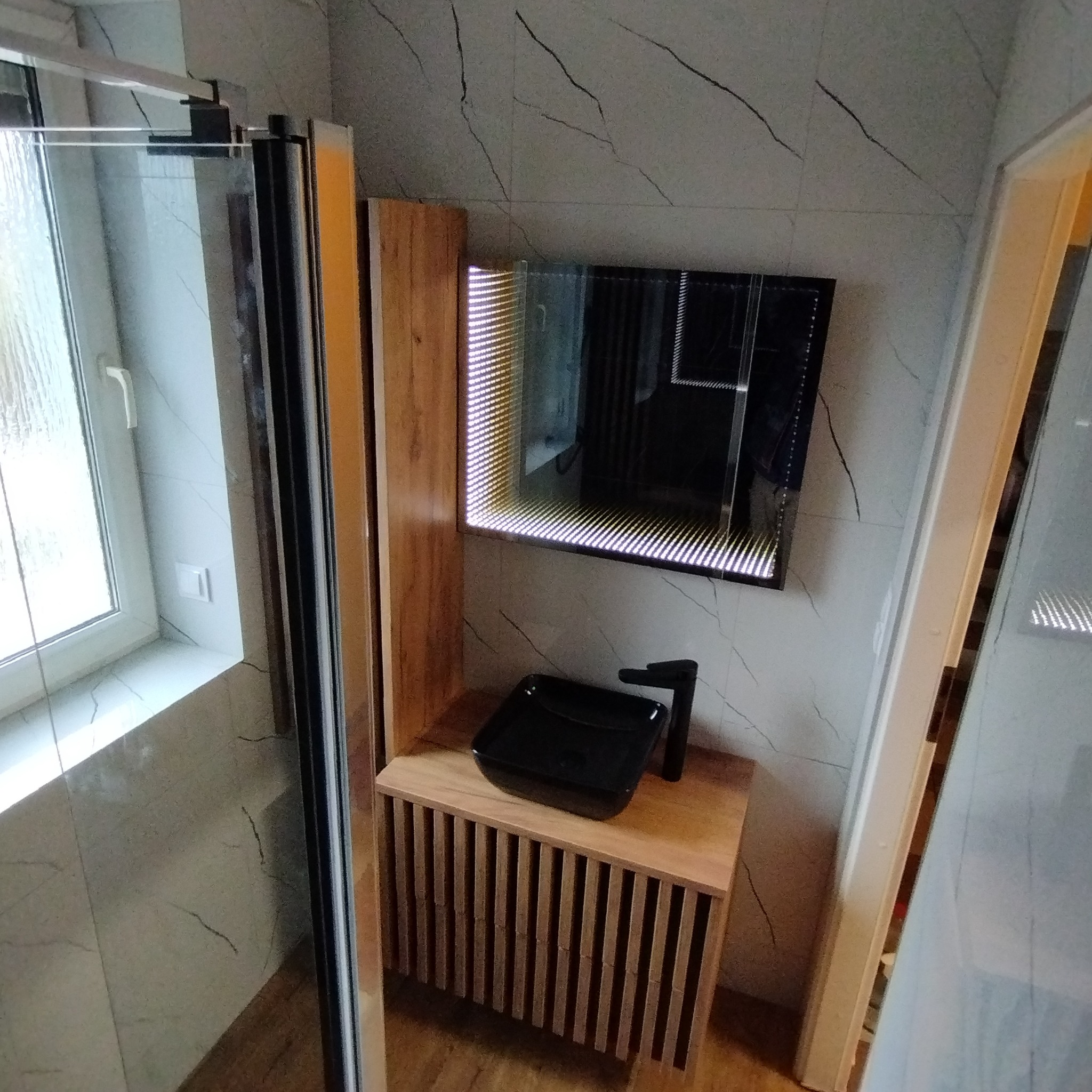 Nowoczesna łazienka z kabiną prysznicową, drewnianą szafką umywalkową z czarną nablatową umywalką i lustrem z efektem nieskończoności, wykończona płytkami imitującymi marmur.