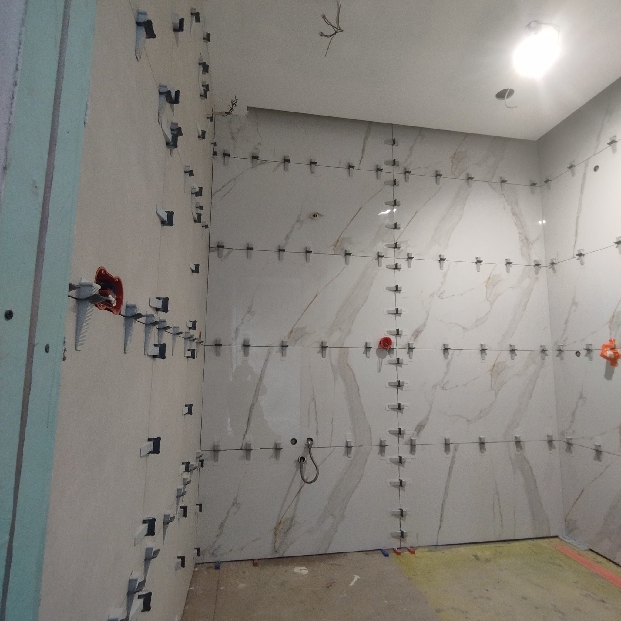 Ściany w trakcie układania płytek imitujących marmur, z użyciem systemu poziomowania płytek, widoczne przewody elektryczne wystające ze ściany.