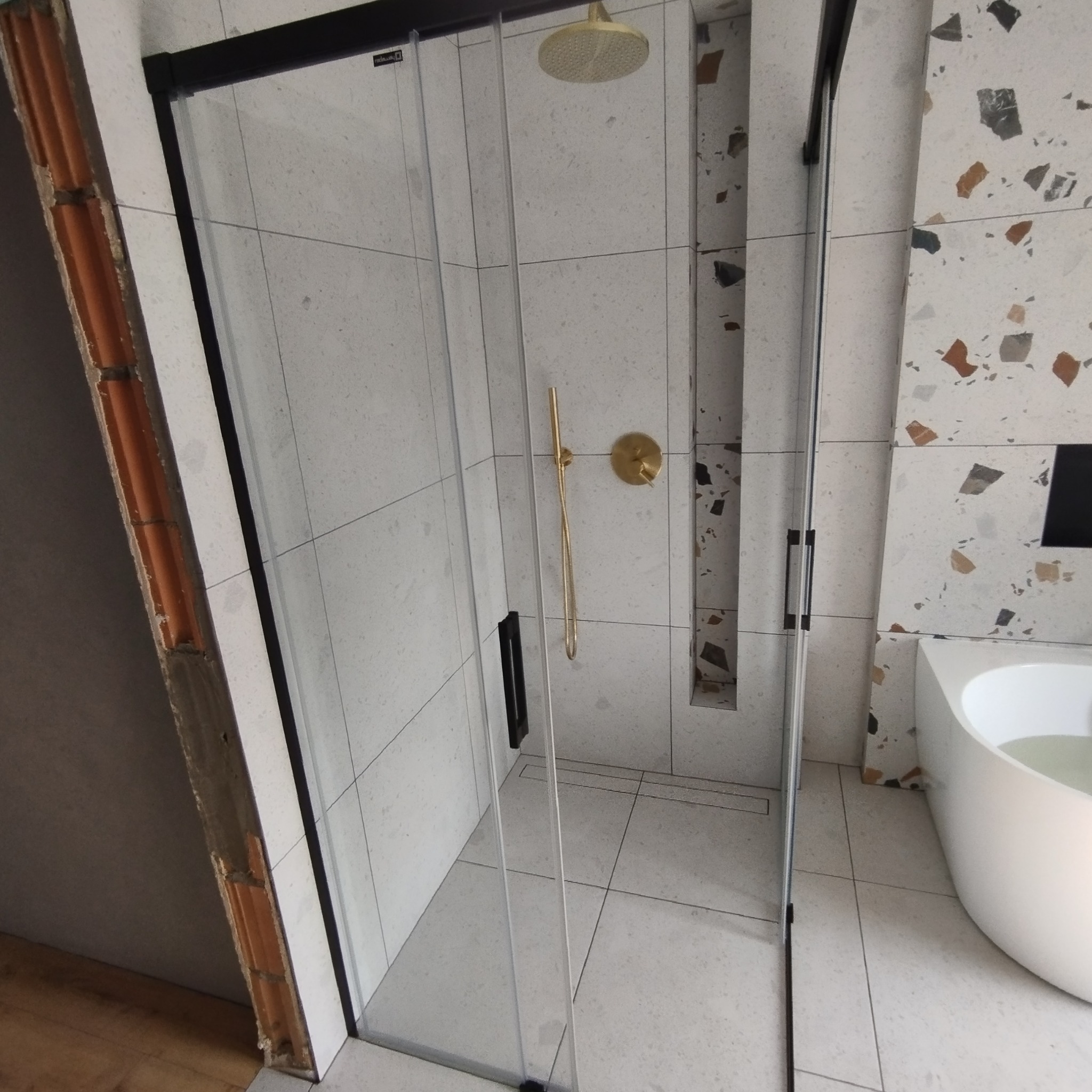 Kabina prysznicowa z czarnymi profilami i złotą armaturą na tle ściany z mozaiką lastryko, widoczny fragment niezakończonego obrzeża ściany z cegły