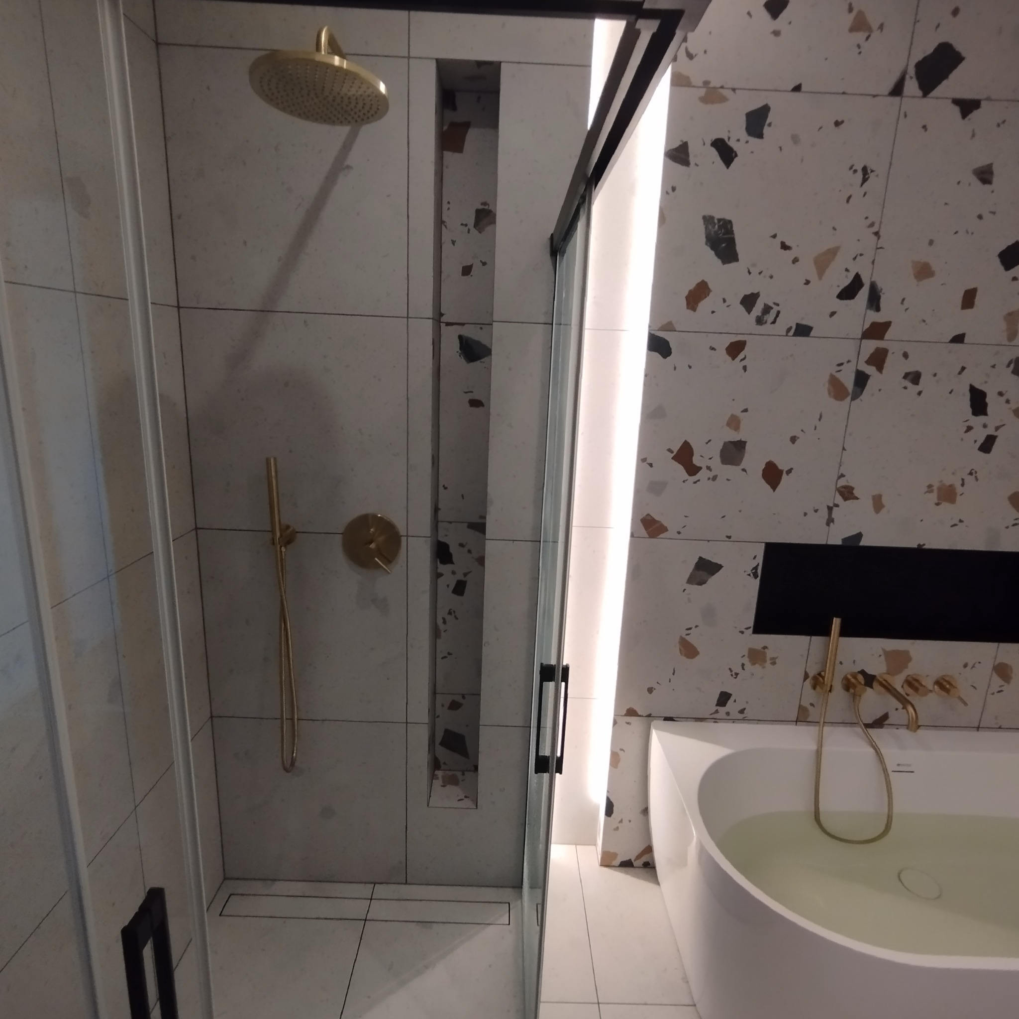 Nowoczesna łazienka z kabiną prysznicową typu walk-in, złotą armaturą, płytkami imitującymi lastryko i wnęką w ścianie.