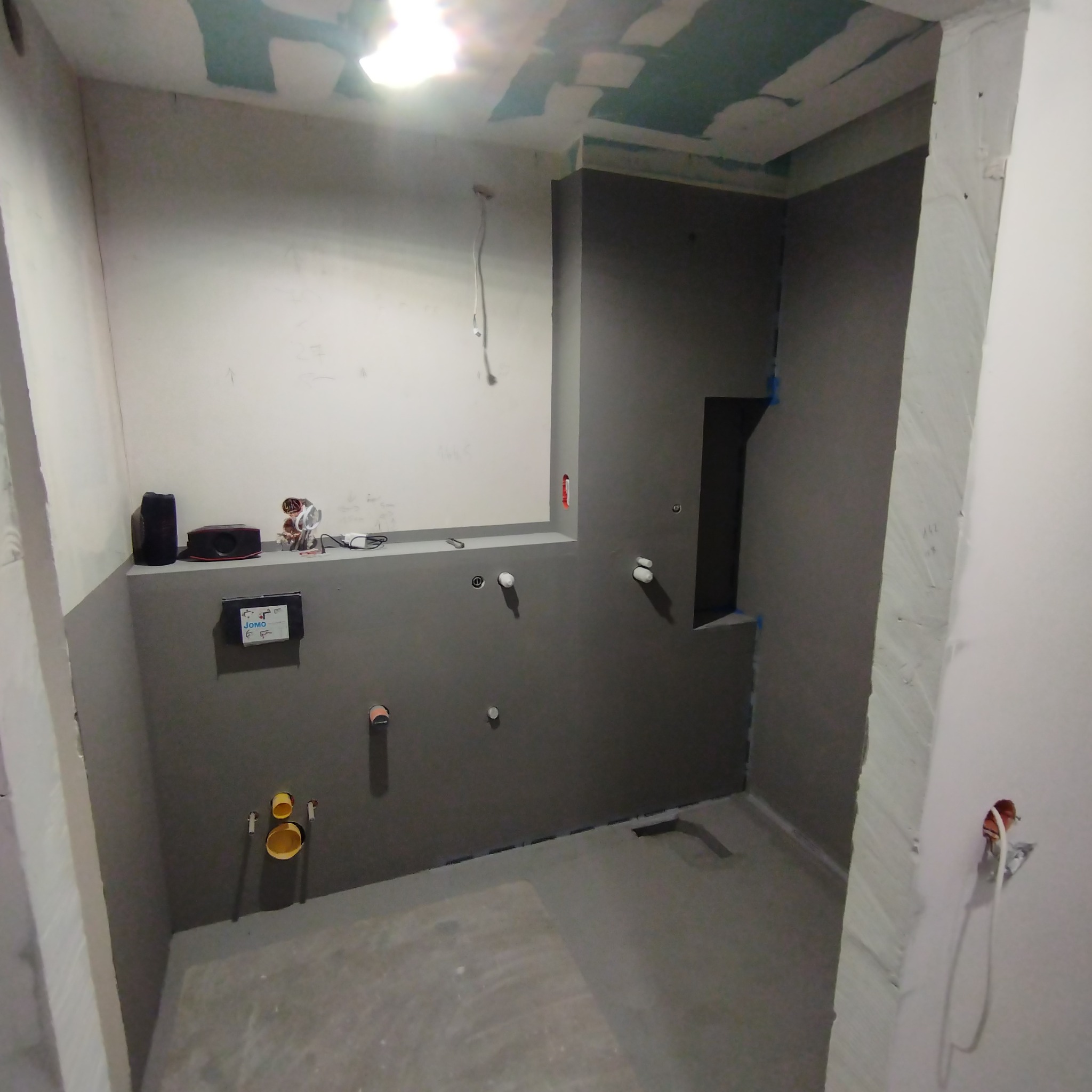 Surowa łazienka w trakcie przygotowania do wykończenia, widoczne instalacje hydrauliczne i elektryczne, ściany częściowo pokryte szarą hydroizolacją, sufit z widocznymi śladami po pracach...