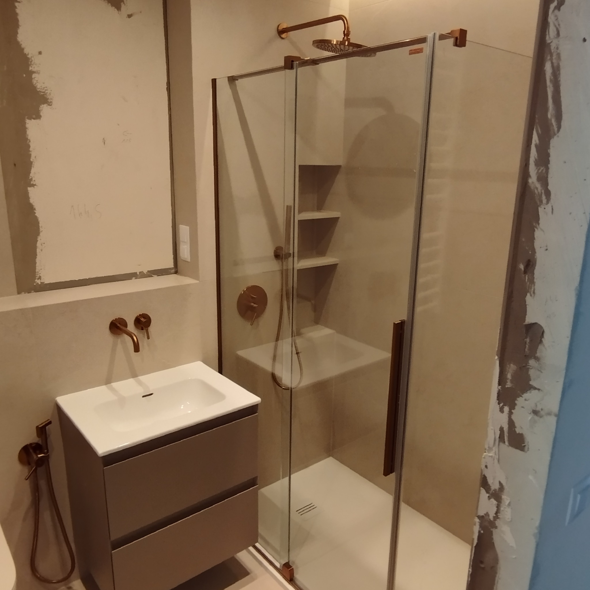 Nowoczesna łazienka w trakcie wykańczania, widoczna kabina prysznicowa z armaturą w kolorze miedzi, umywalka z szafką i bidetką, ściany częściowo przygotowane do malowania lub położenia płytek.