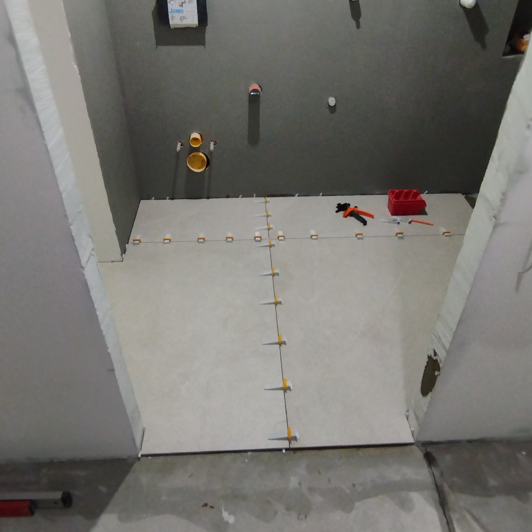 Układanie jasnych płytek podłogowych w łazience z użyciem systemu poziomowania płytek, widoczne narzędzia glazurnicze i przygotowane otwory instalacyjne w szarej ścianie.