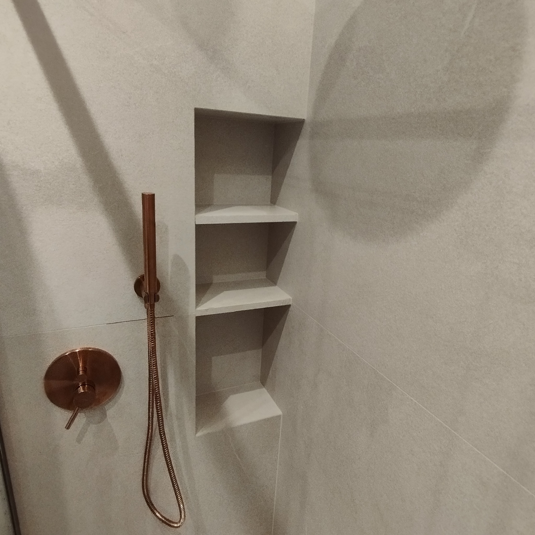 Szare płytki w łazience z wnęką z trzema półkami i miedzianą armaturą prysznicową.