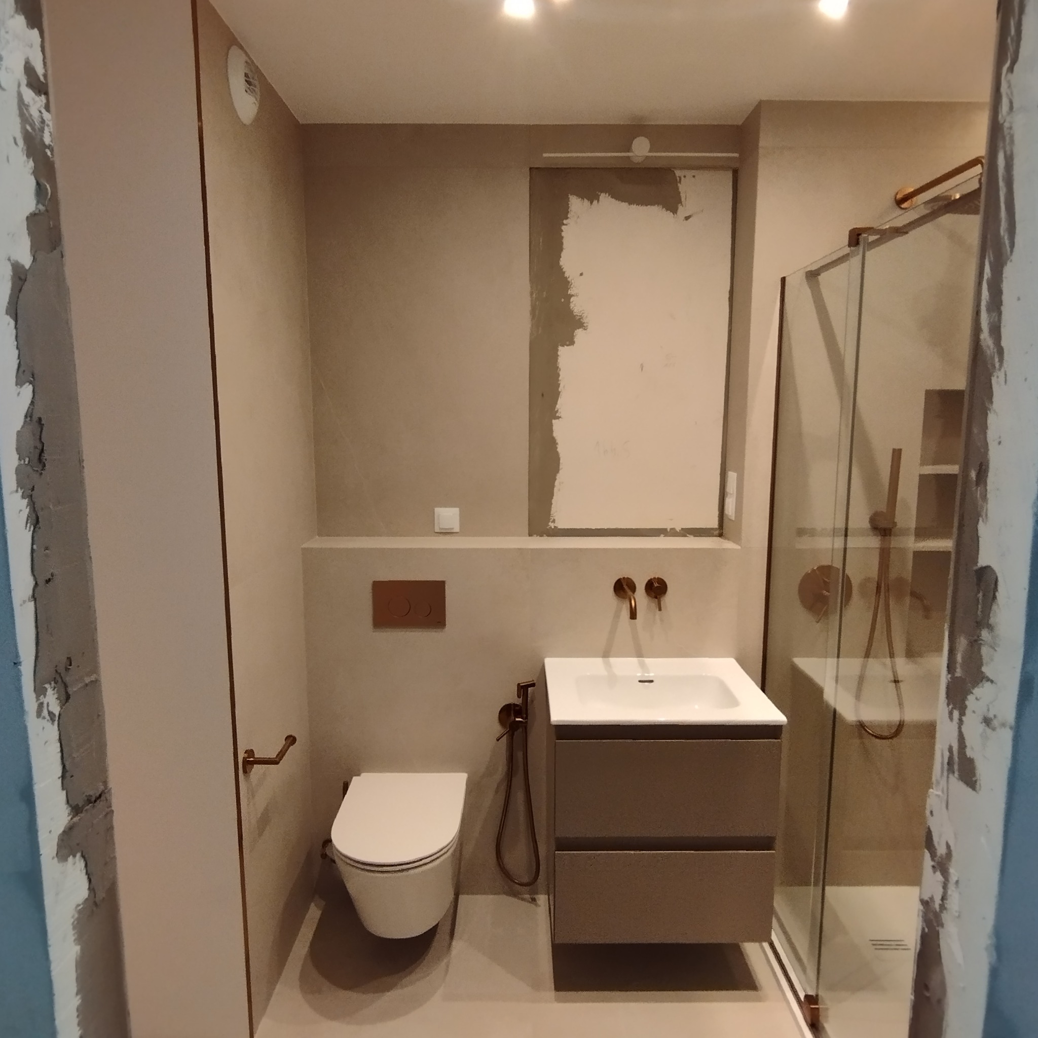 Nowoczesna łazienka w trakcie remontu z wiszącą toaletą, szafką z umywalką i kabiną prysznicową z elementami w kolorze miedzi. Fragment ściany z niedokończonym wykończeniem.