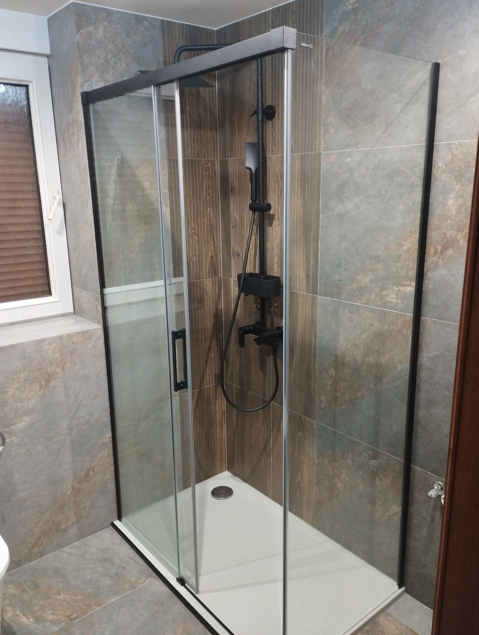 Nowoczesna kabina prysznicowa z czarnymi profilami i deszczownicą, obłożona płytkami imitującymi drewno i kamień, w jasnej łazience z oknem.