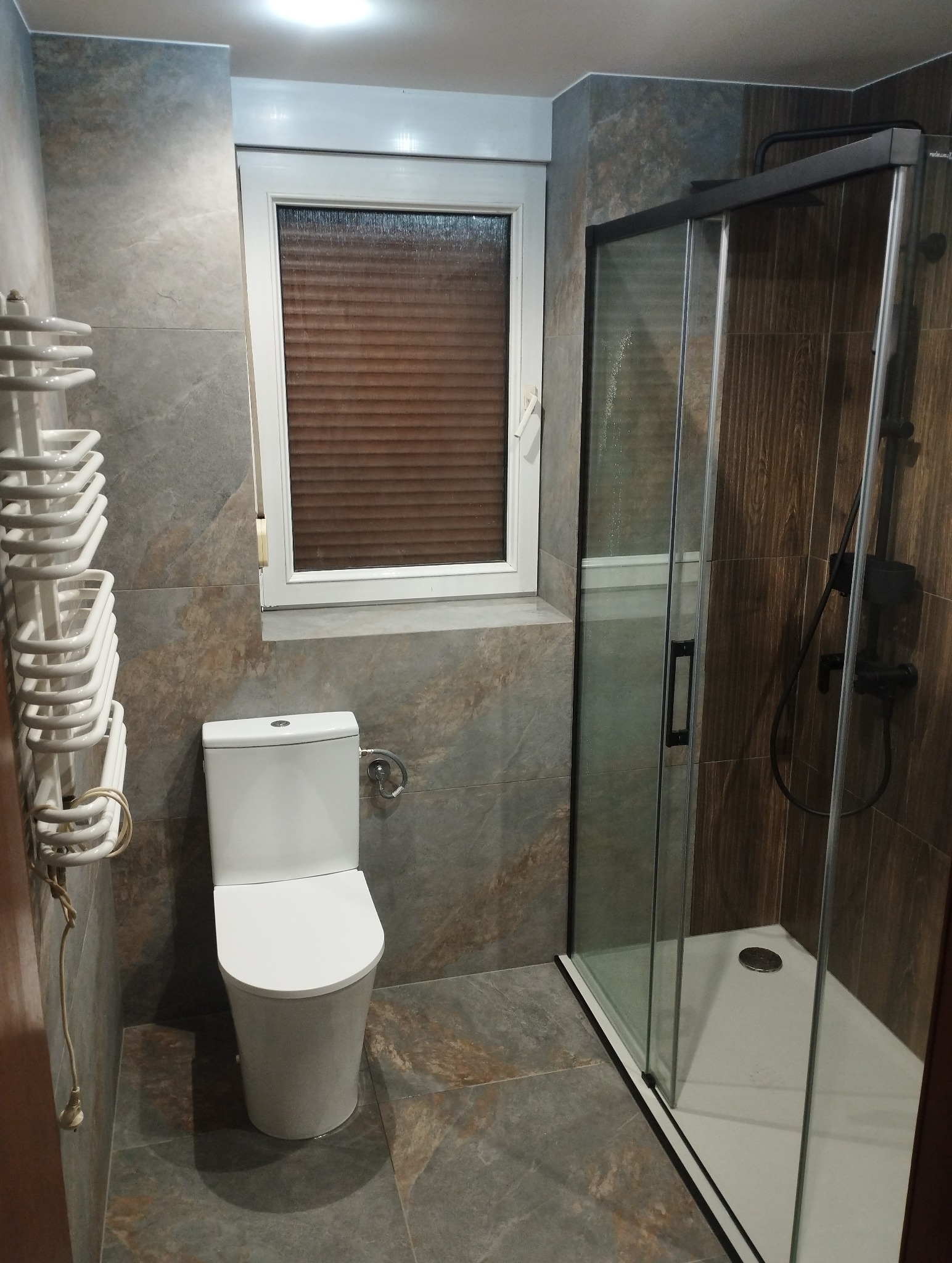 Nowoczesna łazienka z szarymi płytkami imitującymi kamień, kabiną prysznicową z czarnymi ramami i białym WC, oświetlona przez okno z roletą.