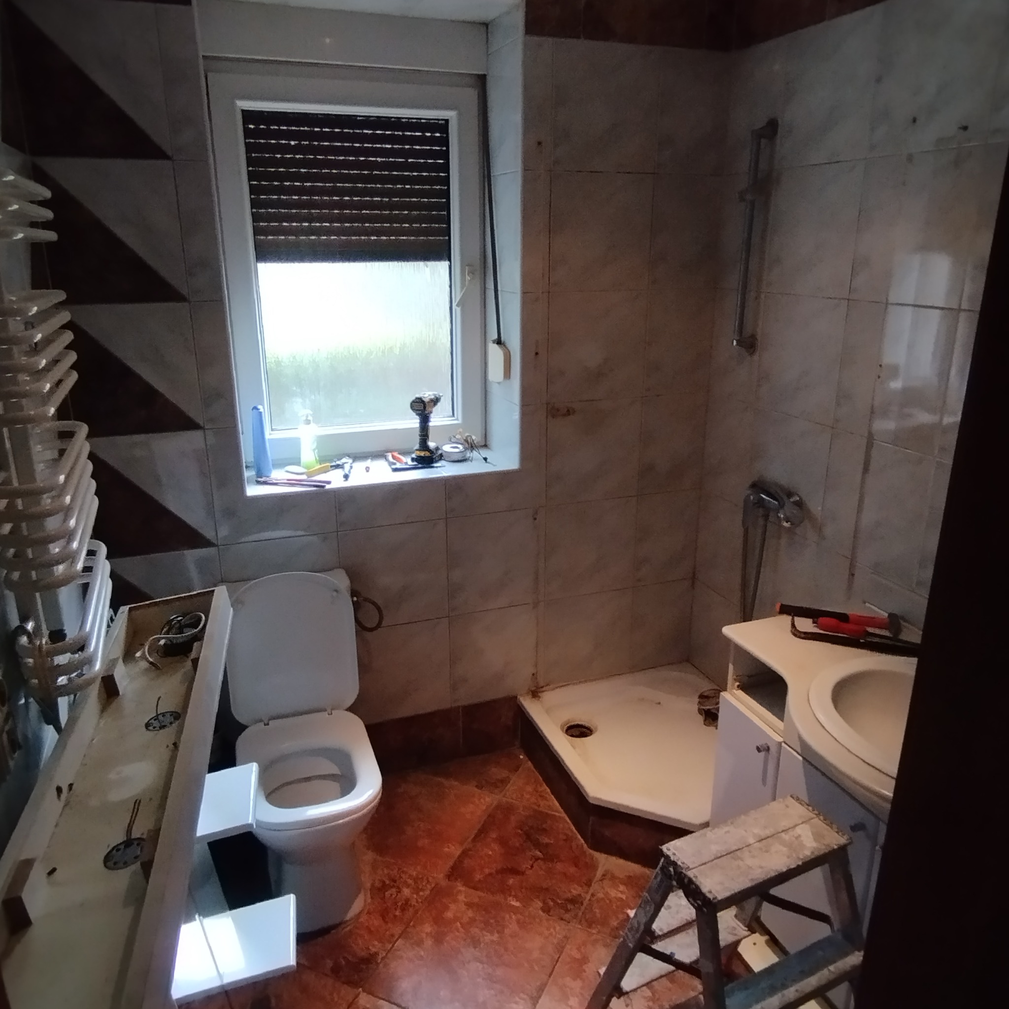 Łazienka w trakcie remontu: widoczny sedes, brodzik, drabina, narzędzia na parapecie. Płytki w odcieniach brązu i beżu. Grzejnik drabinkowy częściowo zamontowany.