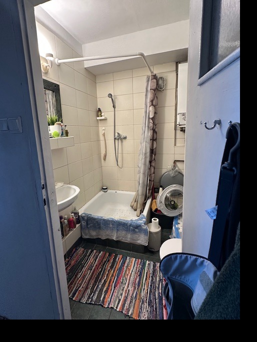 Wnętrze małej łazienki z wanną obudowaną niebieską tkaniną, pralką i kolorowym dywanikiem. Widok zza uchylonych drzwi.